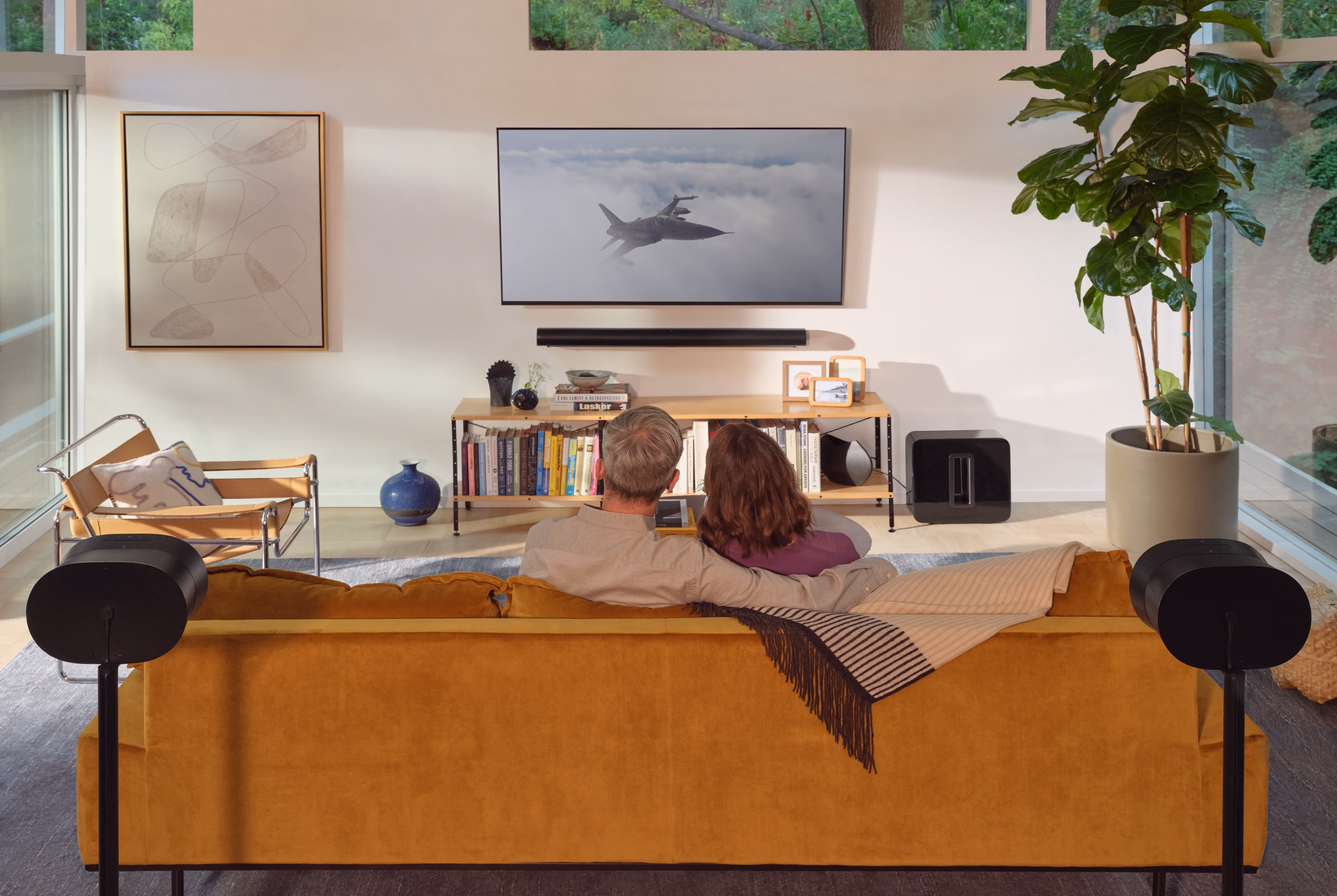 Modern living room with a flat-screen TV, large speakers, and a leather sofa. Natural light from a large window and a split air conditioning unit above enhance the ambiance.